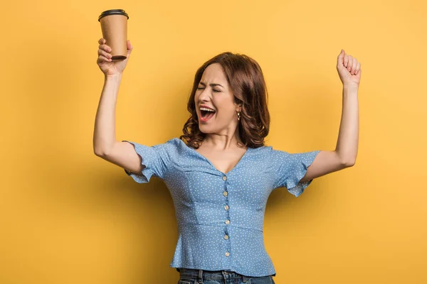 Excited girl showing winner gesture while holding paper cup on yellow background — Stock Photo