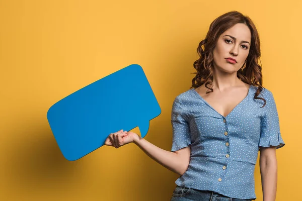 Displeased girl looking at camera while holding speech bubble on yellow background — Stock Photo