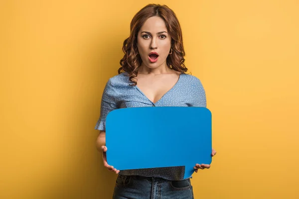 Shocked girl looking at camera while holding speech bubble on yellow background — Stock Photo