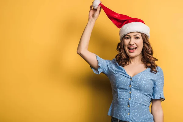 Happy girl touching santa hat while looking at camera on yellow background — Stock Photo