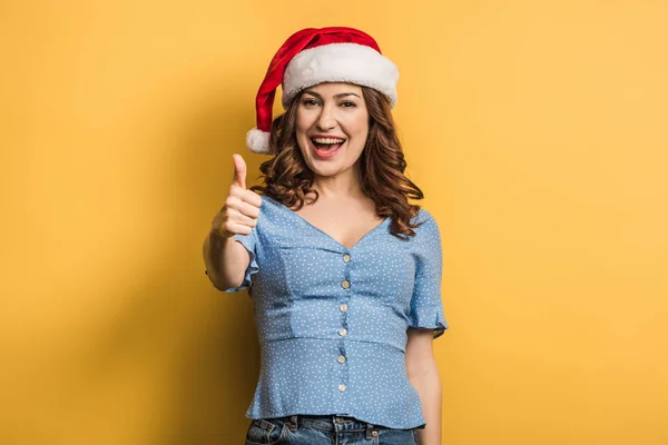 Cheerful girl in santa hat showing thumb up on yellow background — Stock Photo