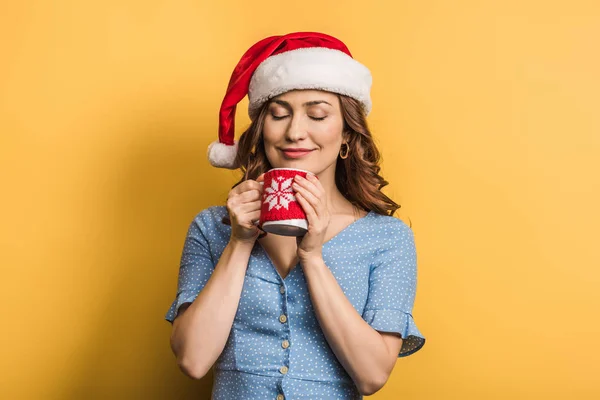 Joyful girl in santa hat holding cup of warm drink with closed eyes on yellow background — Stock Photo