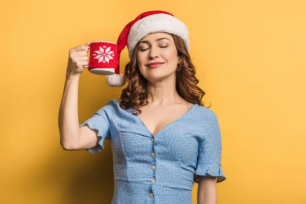 Compiaciuta ragazza in cappello di Santa in possesso di tazza di bevanda calda con gli occhi chiusi su sfondo giallo — Foto stock