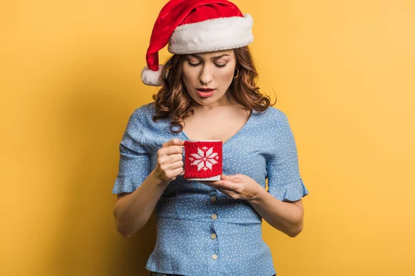 Незадоволена дівчина в капелюсі Санта дивиться на чашку на жовтому фоні — стокове фото