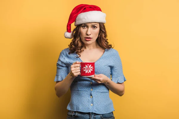 Displeased girl in santa hat holding cup with warming drink on yellow background — Stock Photo