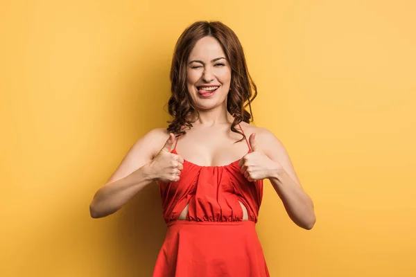 Cheerful girl showing thumbs up, winking and sticking out tongue on yellow background — Stock Photo