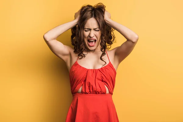 Mujer joven enojada gritando de pie con los ojos cerrados y tocando el pelo en el fondo amarillo - foto de stock