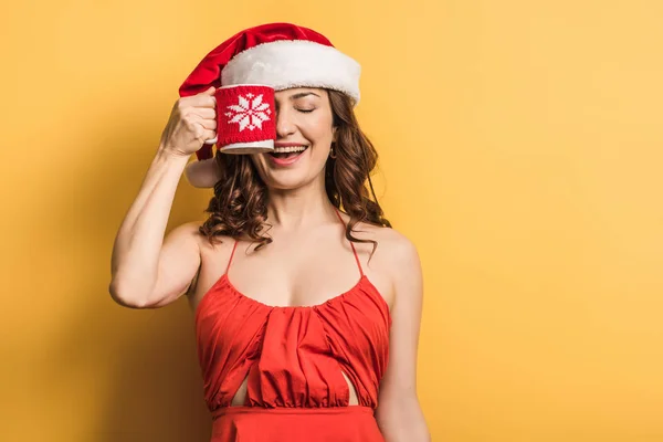 Smiling girl in santa hat covering eye with cup on yellow background — Stock Photo