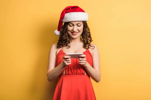 Весела дівчина в капелюсі Санта на смартфоні на жовтому фоні — стокове фото
