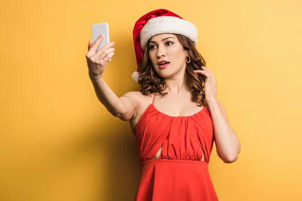 Elegant woman in santa hat taking selfie on smartphone on yellow background — Stock Photo