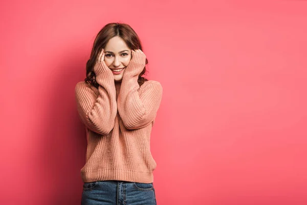 Sonriente chica cogida de la mano cerca de la cara mientras mira a la cámara en el fondo rosa - foto de stock