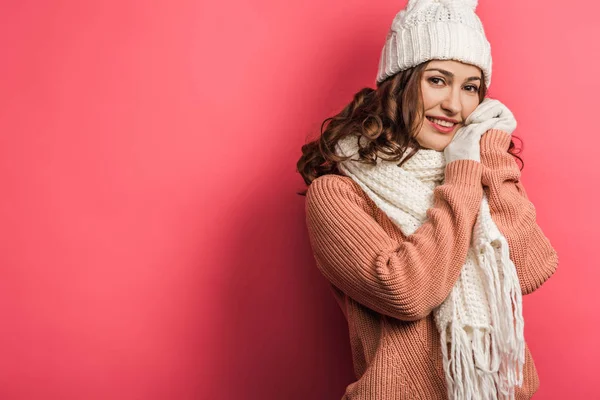 Chica fría y sonriente en sombrero caliente y bufanda de pie con las manos apretadas sobre fondo rosa - foto de stock