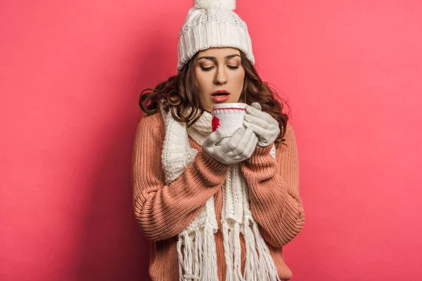 Cold girl in warm hat and scarf holding cup with warming drink on pink background — Stock Photo