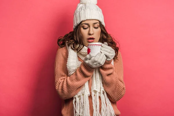 Fille froide en chapeau chaud et écharpe tenant tasse avec boisson chauffante sur fond rose — Photo de stock