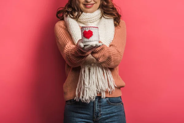 Cropped view of smiling girl in warm scarf holding cup with heart symbol on pink background — Stock Photo