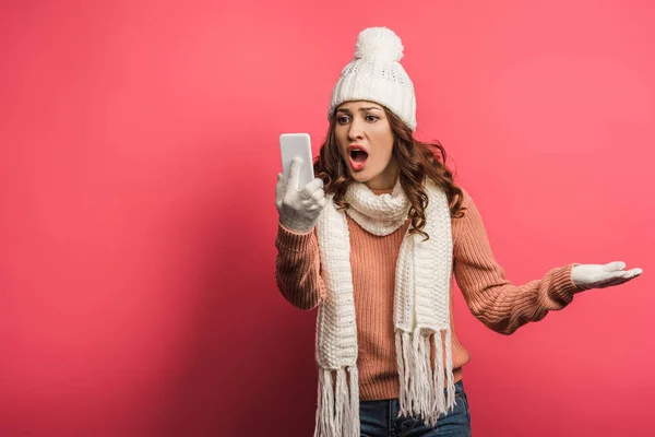 Angry girl in warm hat and scarf screaming during video call on smartphone on pink background — Stock Photo