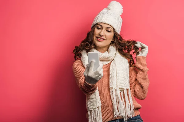 Flirty girl in warm hat and scarf touching hair during video call on smartphone on pink background — Stock Photo
