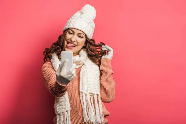 Tricky girl in warm hat and scarf sticking out tongue during video call on smartphone on pink background — Stock Photo