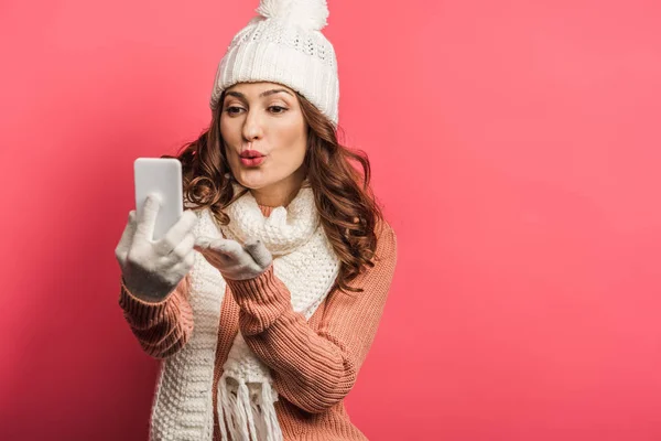 Positive girl in warm hat and scarf sending air kiss during video call on smartphone on pink background — Stock Photo