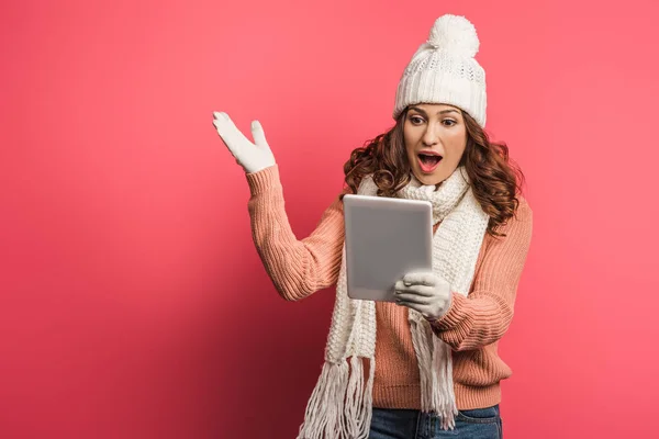 Chica sorprendida en sombrero caliente y bufanda mirando tableta digital sobre fondo rosa - foto de stock