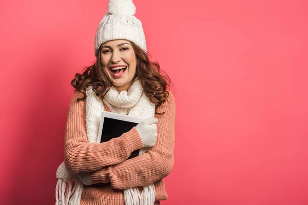 Cheerful girl in warm hat and scarf holding digital tablet on pink background — Stock Photo