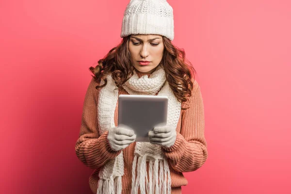 Chica concentrada en sombrero caliente y bufanda usando tableta digital sobre fondo rosa - foto de stock