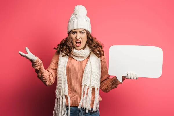 Irritated girl in warm hat and scarf holding speech bubble on pink background — Stock Photo