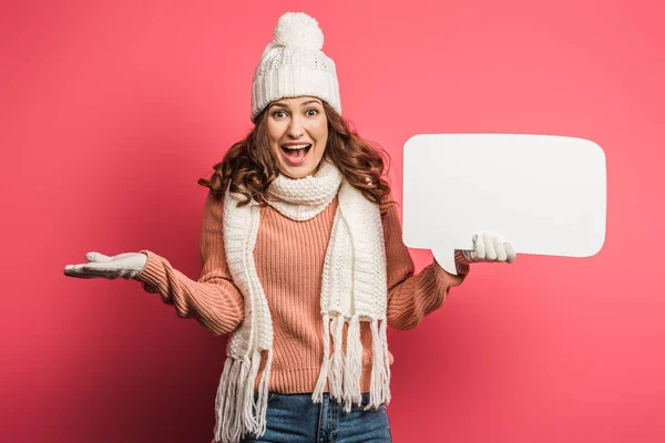 Cheerful girl in warm hat and scarf holding speech bubble on pink background — Stock Photo