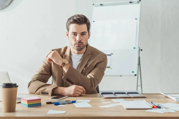 Gutaussehender UX-Designer blickt in die Kamera, während er am Tisch mit Webseitenskizzen sitzt — Stockfoto