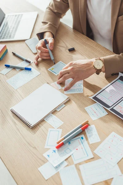 Vue recadrée du concepteur développant des mises en page d'applications à côté des croquis de wireframe mobiles sur la table — Photo de stock