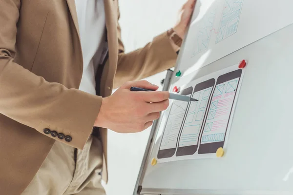 Vista recortada del diseñador haciendo notas en bocetos de interfaz de aplicación en pizarra en la oficina - foto de stock