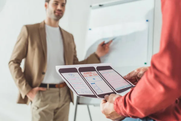 Selective focus of designer holding ux mobile application layout with colleagues beside whiteboard at background — Stock Photo