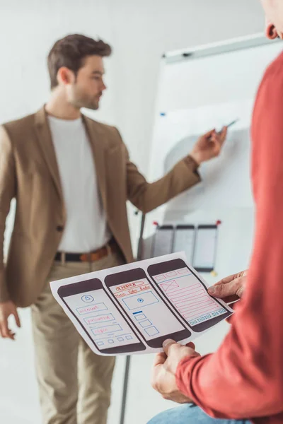 Selective focus of creative designer holding mobile website sketches while colleague whiting on whiteboard in office — Stock Photo