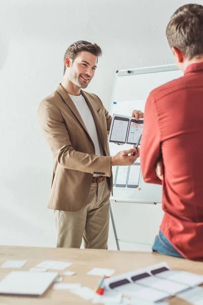 Enfoque selectivo del diseñador sonriente que muestra las pantallas del desarrollo del sitio web móvil sensible al colega en la oficina - foto de stock