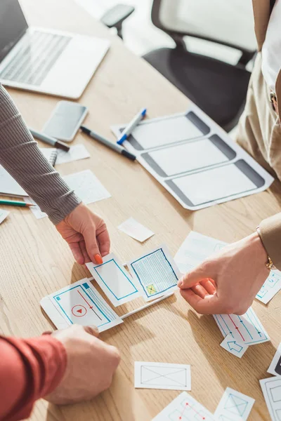 Vista recortada de los diseñadores ux con bocetos web y prototipo móvil trabajando en la mesa - foto de stock