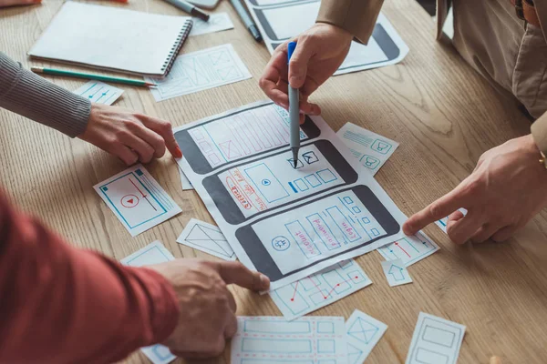 Vista recortada de diseñadores que trabajan con bocetos de aplicaciones y plantilla de sitio web móvil en la mesa - foto de stock