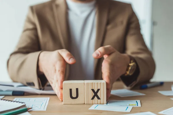 Vista cortada de designer segurando cubos de madeira com letras ux na mesa com layouts — Fotografia de Stock