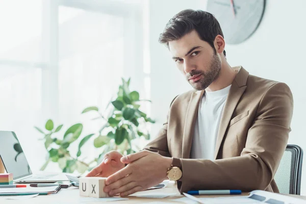 Schöner Designer blickt in die Kamera, während er Würfel mit UX-Buchstaben am Arbeitstisch hält — Stockfoto