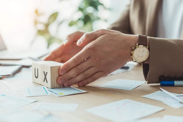 Vista cortada de designer segurando cubos de madeira com letras ux ao lado de esboços de aplicativos na mesa — Fotografia de Stock