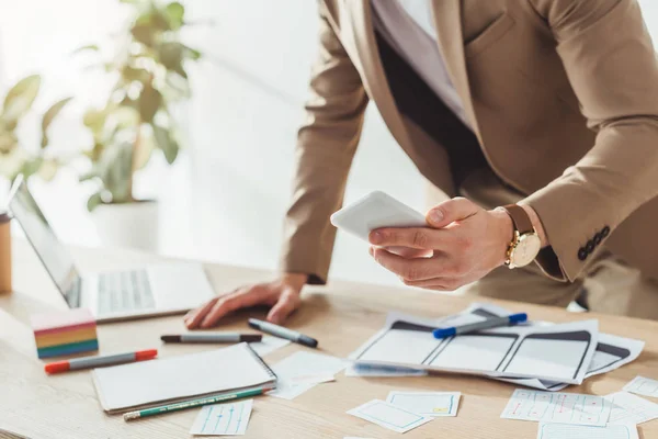 Vista recortada del diseñador ux utilizando el teléfono inteligente por el sitio web bocetos de marco de alambre en la mesa — Stock Photo