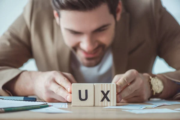 Enfoque selectivo del diseñador creativo sonriente sosteniendo cubos con letras ux en la mesa con diseños — Stock Photo