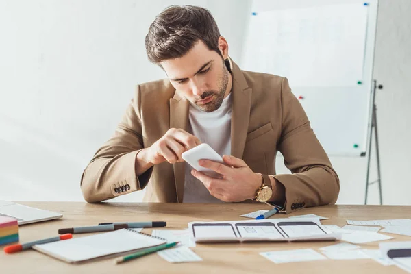 Selektiver Fokus des gutaussehenden UX-Designers per Smartphone neben App-Skizzen auf dem Tisch — Stockfoto