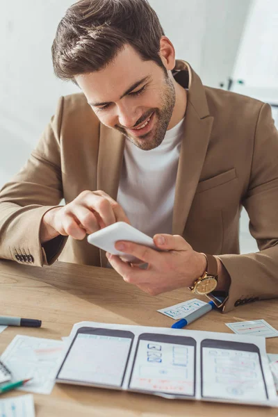 Selective focus of smiling designer using smartphone beside wireframe sketches of mobile website on table — Stock Photo