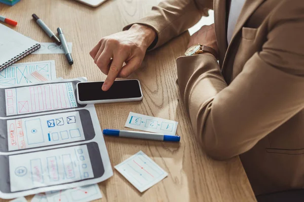 Vista recortada do designer usando o smartphone enquanto desenvolve o design da experiência do usuário na mesa — Fotografia de Stock