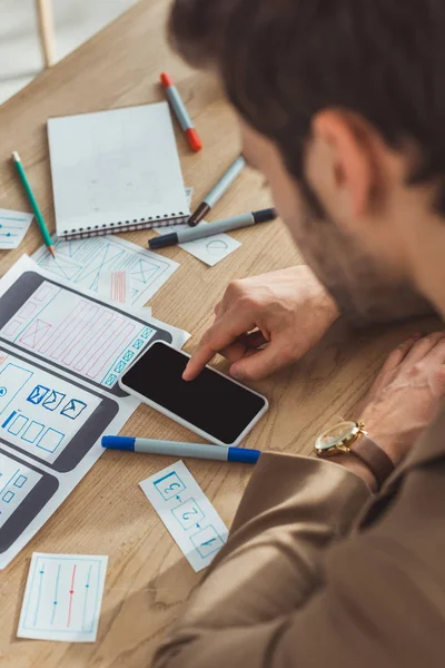 Enfoque selectivo del diseñador creativo utilizando el teléfono inteligente por bocetos web y diseños en la mesa - foto de stock