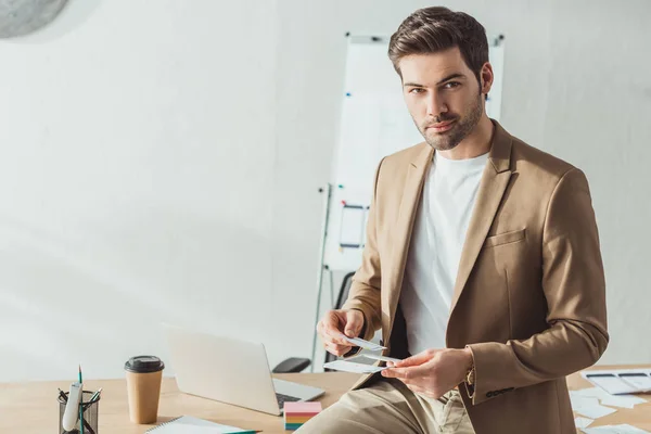 Designer blickt auf Kamera, während er Layouts und mobile Website-Vorlage im Büro hält — Stockfoto