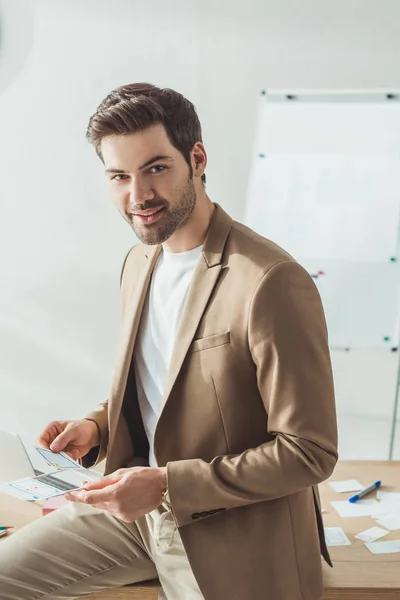 Guapo diseñador mirando a la cámara mientras sostiene bocetos por mesa - foto de stock