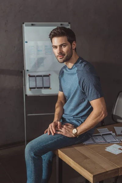 Gutaussehender Designer blickt auf Kamera, während er auf einem Tisch mit Grundrissen sitzt — Stockfoto