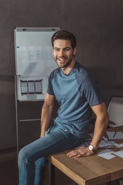 Beau ux designer souriant à la caméra tout en étant assis sur la table avec des croquis — Photo de stock