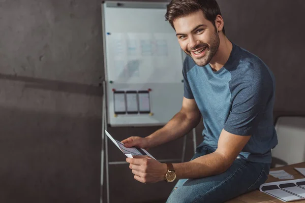 Designer sorridente alla fotocamera mentre tiene modello di sito web mobile in studio di progettazione — Foto stock